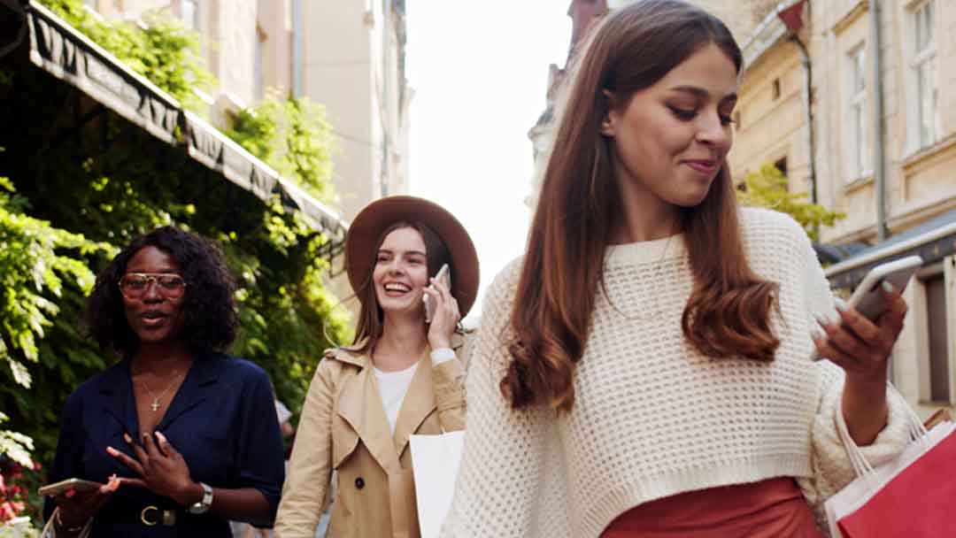 Three women shopping local businesses outdoors, using smartphones and laughing, symbolizing internet marketing and local digital marketing success.